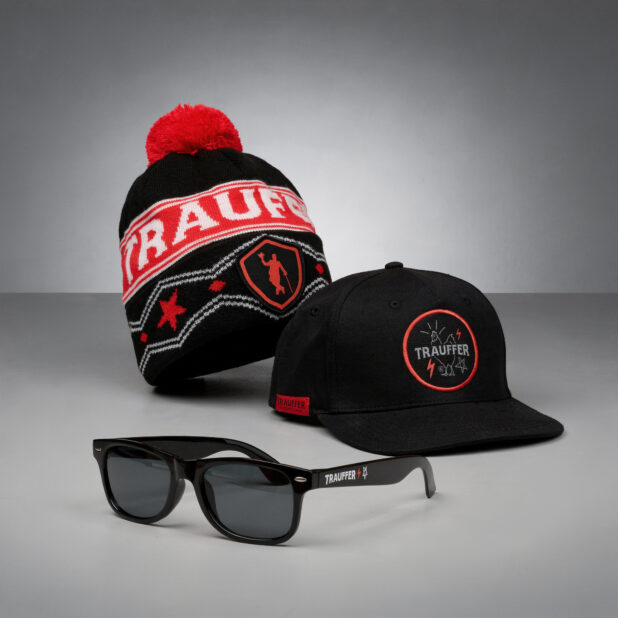 Eine schwarz-rote Strickmütze, eine schwarze Cap und schwarze Sonnenbrillen aus der Trauffer Alpentainer Branded Merch Kollektion, alle mit dem Trauffer-Logo, präsentiert auf einer grauen Fläche vor grauem Hintergrund